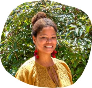 Nikki smiling in a yellow tunic and red earrings in front of green plants